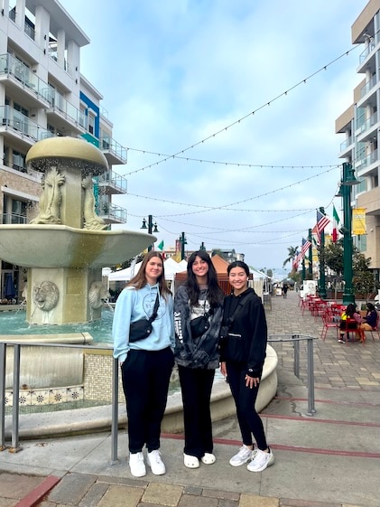 Three young girls posing in front of an outdoor fountain at the farmer's market in Little Italy, Carlsbad, California. They stand beween two rows of buildings with blue sky and hanging lights in the background.